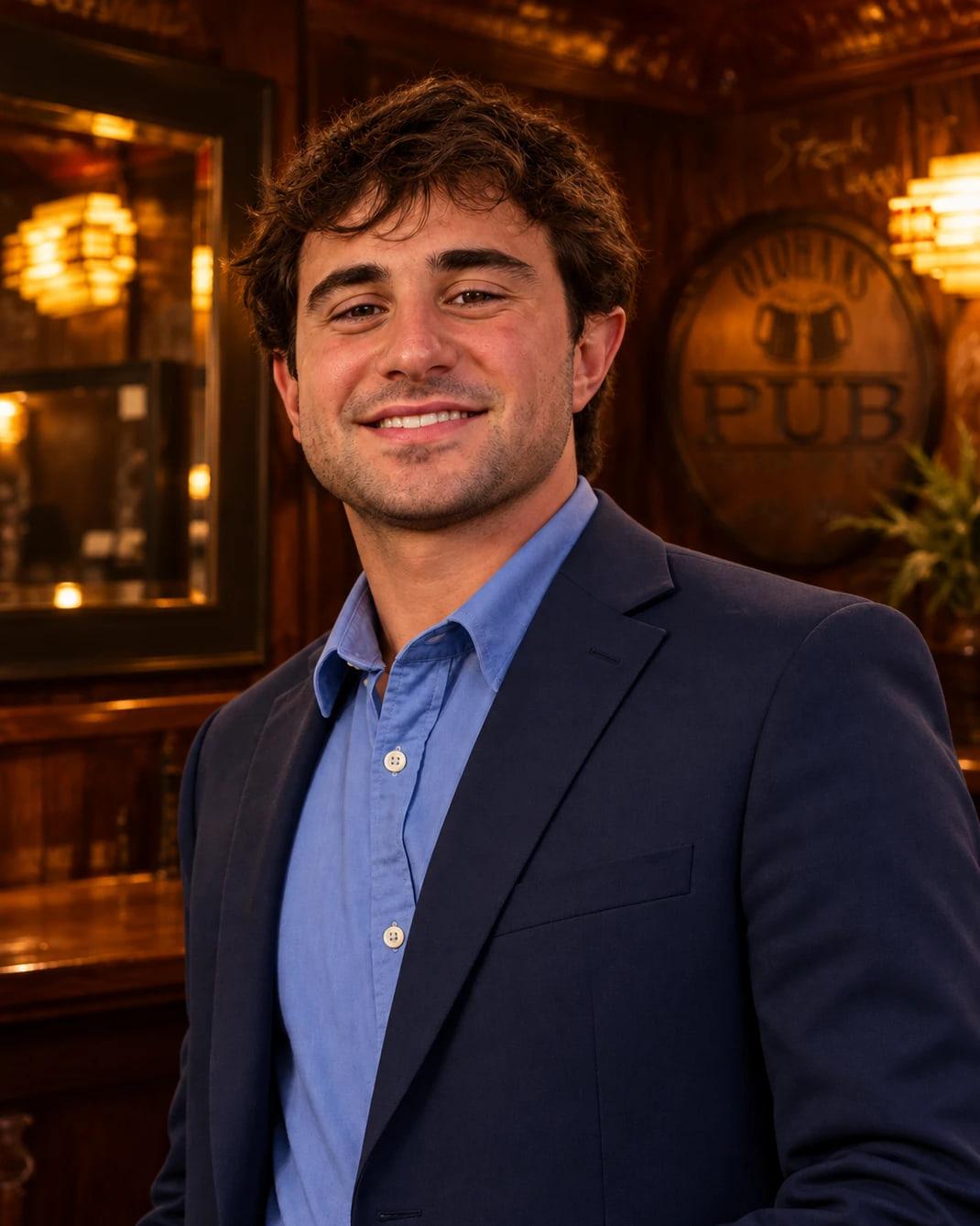 Cecco Logrippo smiling in a navy blazer and blue shirt, in front of a wood-paneled wall with the Olohan’s Pub sign and stained-glass lamps.