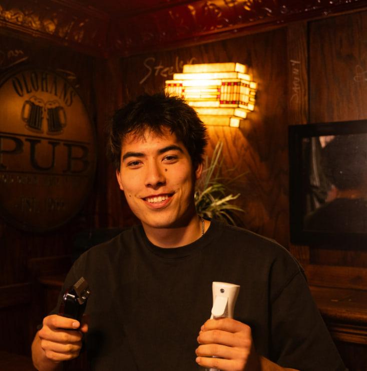 William Olohan smiling with clippers and a spray bottle in hand, in front of a wood-paneled wall and a glowing stained-glass lamp.